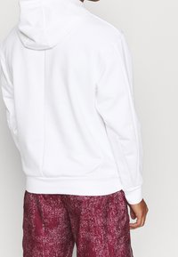 Sweat à capuche blanc avec une texture lisse, présentant un détail de couture au dos. Associé à un short bordeaux à motif.