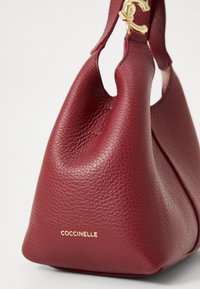 Burgundy leather handbag with a textured finish, featuring a rounded shape and gold hardware accent. Logo displayed in gold on the front.