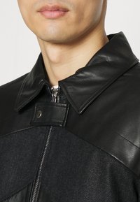 Veste en cuir noir et en laine chevron avec un col montant, fermeture éclair en métal et détail de fermeture à pression au niveau du cou. Superposition texturée visible.