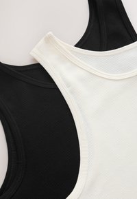 White and black ribbed tank tops laid overlapping on a light surface, showing stitching and fabric texture clearly.