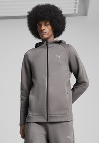 Grå lynlås-hættetrøje lavet af blødt materiale med slim fit, med et lille hvidt logo på brystet og sorte lynlåsdetaljer.