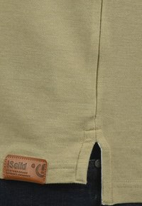 Oliven grøn bomuld t-shirt med en tekstureret overflade, korte sprækker i siderne og et brunt lædermærke med brandnavnet.