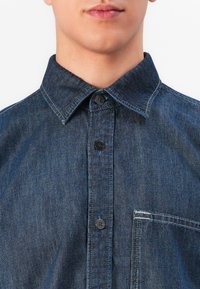 Man wearing dark blue denim button-up shirt with black buttons and a stitched chest pocket, shown from neck to mid-torso.