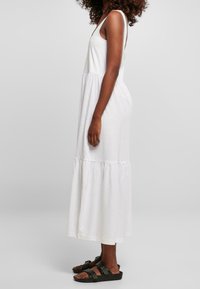 Biała bawełniana sukienka maxi z bezrękawowym krojem, wielowarstwowym dołem i luźnym fasonem. W połączeniu z ciemnymi sandałami. Widok z boku.