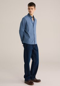 Camisa de manga larga color azul claro con diseño de botones, combinada con jeans azul oscuro. La camisa presenta un pequeño logo, textura suave y un corte ajustado.