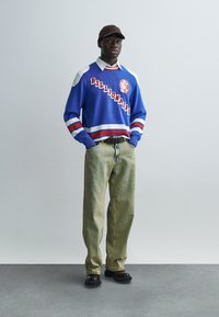 Μπλε πλεκτό πουλόβερ με κόκκινες λεπτομέρειες και το κείμενο «Billionaire», συνδυασμένο με ανοιχτοπράσινα τζιν παντελόνια φαρδιάς γραμμής και μαύρα παπούτσια. Φορά καφέ καπέλο.