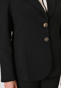 Blazer nero su misura con tre bottoni metallici, cuciture laterali e una texture liscia. Progettato con una vestibilità strutturata e colletto a rever.