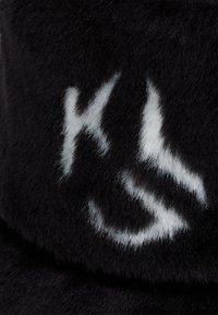 Črna krznena kapa z mehkobo, z izrazitim belim napisom, ki spominja na črki "K" in "S" spredaj, kar ustvarja močan kontrast.