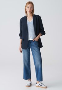 Blazer marine sur un top rayé blanc, associé à un jean bleu ample et des baskets blanches avec des accents marins. Tissu lisse et coupe moderne.