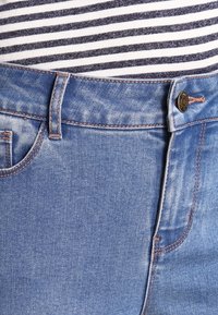 Ljusblå denimjeans med dragkedja och knappstängning, med orange sömmar, fem fickor och normal midja. Randig topp delvis synlig.