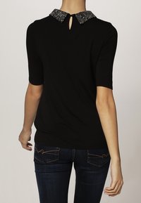 Kvinna klädd i en svart kortärmad topp med en glittrig pärlkrage, detalj med nyckelhål på baksidan, samt mörkblå jeans med sydda bakfickor.