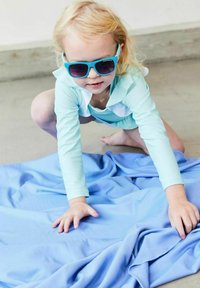Light blue long-sleeve swim shirt, white accents, and blue sunglasses on a child kneeling on soft blue fabric. Textured, smooth surface.