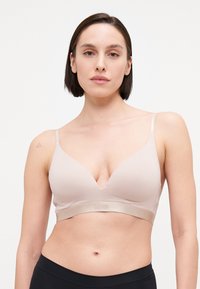 Lys beige bralette med dyp utringning, justerbare stropper og en myk stropp med embossed merkevare. Glatt tekstur.