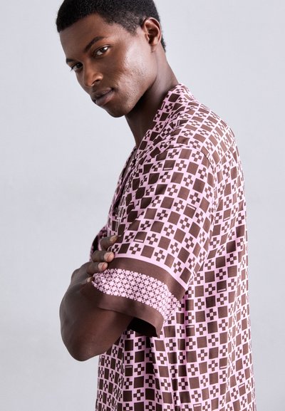 Un jeune homme les bras croisés portant une chemise à manches courtes rose et brune à motifs devant un fond clair.