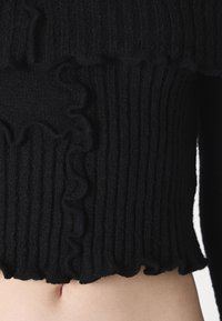 Pull en tricot noir avec une texture côtelée, orné de volants le long de l'ourlet et des coutures latérales, coupe courte.