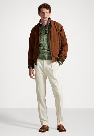 Braune Wildleder-Bomberjacke über einem grünen Kabelstrickpullover, cremefarbene Hose und braune Loafers; Accessoires umfassen Sonnenbrille und ein Hemd mit Kragen.
