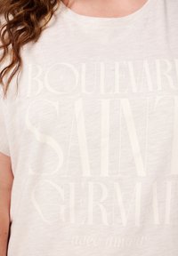 T-shirt en coton beige clair avec un grand texte blanc en relief portant la mention "BOULEVARD SAINT GERMAIN avec amour." Manches courtes et coupe décontractée.