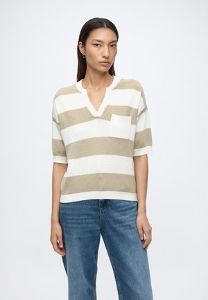 Femme portant un pull à manches courtes à larges rayures beige et blanches avec une poche poitrine et un jean bleu, debout devant un fond uni.