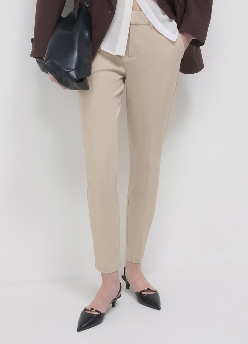 Pantalon beige sur mesure avec une coupe slim, des poches latérales et une texture lisse, associé à des chaussures à bride arrière noires à bouts pointus.