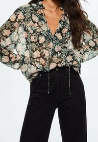 Femme portant un chemisier transparent à manches longues avec imprimé floral et volants, ainsi qu'un pantalon noir taille haute, se tenant debout les mains sur les hanches.