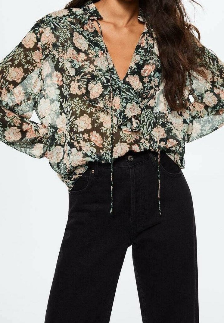 Femme portant un chemisier transparent à manches longues avec imprimé floral et volants, ainsi qu'un pantalon noir taille haute, se tenant debout les mains sur les hanches.