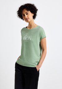 Světle zelené tričko s krátkým rukávem vyrobené z měkké tkaniny, s bílým grafickým potiskem „BON“ na přední straně, v kombinaci s černými kalhotami.