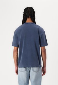 Polo majica iz osnovnega bombaža v mornarsko modri barvi, oversize kroja, s klasičnim ovratnikom in kratkimi rokavi, značilna sproščenega kroja in nepravilnim robom, v kombinaciji s svetlimi denim hlačami.