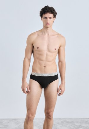 Cuecas pretas de algodão com uma cintura elástica cinzenta que apresenta um logótipo, design ajustado e textura suave. Modelo em pé com os braços ao lado do corpo.