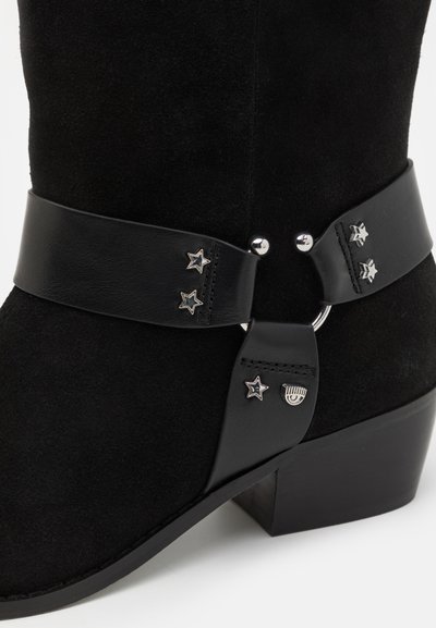 Black suede boot with low heel, featuring decorative black leather straps with silver star and eye-shaped metal studs.