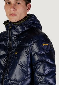 Giacca imbottita blu marino con chiusura a zip, cappuccio e finitura testurizzata. Presenta dettagli gialli e una toppa con logo sulla spalla.