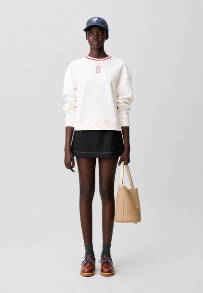 Sweat-shirt blanc avec col rouge, mini-jupe noire, chaussettes grises, chaussures marron avec accents bleus, et sac beige. Casquette marine avec logo.