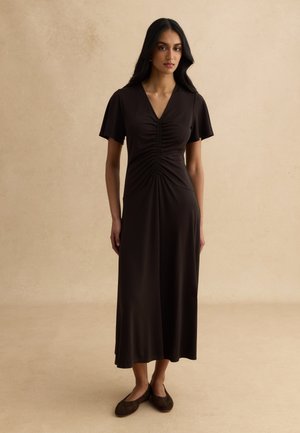 Mujer con el pelo largo y oscuro que lleva un vestido midi negro fruncido con mangas cortas y zapatos planos negros sobre un fondo beige.