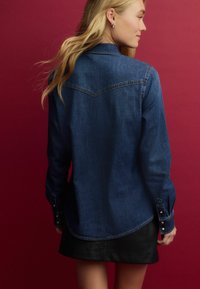 Cămașă din denim albastru închis, cu mâneci lungi, slituri laterale la margine și manșete cu nasturi. Partea din spate are un yoke și o textură subtilă.
