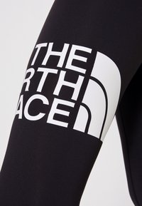 Mustat legginsit, joissa on suuri valkoinen grafiikkapainatus "THE NORTH FACE" -logosta, valmistettu joustavasta, sileästä materiaalista.