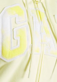 Žlutá mikina s kapucí, na které je velkým výšivkou nápis "GAP" v bílé barvě se žlutými akcenty, s utahovací šňůrkou a měkkou strukturou látky.