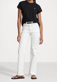 T-shirt noir à manches courtes avec un logo, jeans taille haute blancs, et sandales noires à lanières, se tenant sur un fond gris clair.