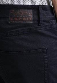 Ciemnoniebieskie dżinsy z widoczną skórzaną metką z napisem "ESPRIT". Cechy obejmują szlufki na pasek, tylnią kieszeń oraz gładką teksturę.