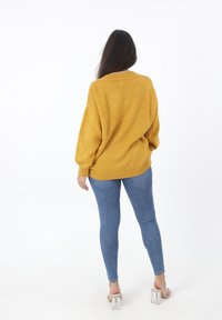 Mosterdgele gebreide trui met een relaxte pasvorm, geribbelde zoom en ingezakte schouders, gecombineerd met blauwe skinny jeans en doorzichtige blokhak sandalen.