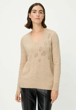 Sweater beige à col en V en tissu tricot doux. Présente des motifs de découpes décoratives d'un côté et des poignets et un ourlet côtelés. Associé à un pantalon noir.