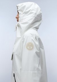 Person wearing white hooded jacket with drawstrings and a beige circular patch on the upper arm, facing left on light gray background.