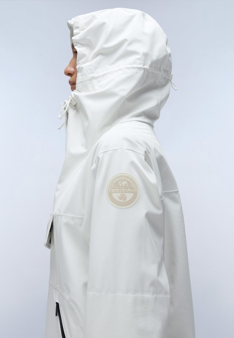 Persoon in een witte hoodie met trekkoorden en een beige ronde patch op de bovenarm, die naar links kijkt op een lichtgrijze achtergrond.