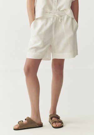 Shorts bianchi in lino con vita elastica e cordoncino. Abbinati a sandali in suede marrone con cinturino a fibbia e suola testurizzata.