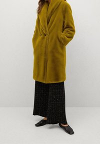 Manteau en fausse fourrure jaune moutarde avec un col châle, deux poches avant et une coupe décontractée, associé à un pantalon noir à jambes larges et des chaussures noires.