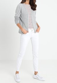 Szary, dziany kardigan z przodem na guziki, połączony z paskowaną czerwoną i białą koszulą, białymi spodniami slim-fit oraz białymi trampkami.