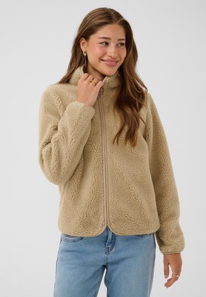 Jeune femme portant une veste polaire beige et un jean bleu, tenant le col de la veste et souriante, debout devant un fond uni.