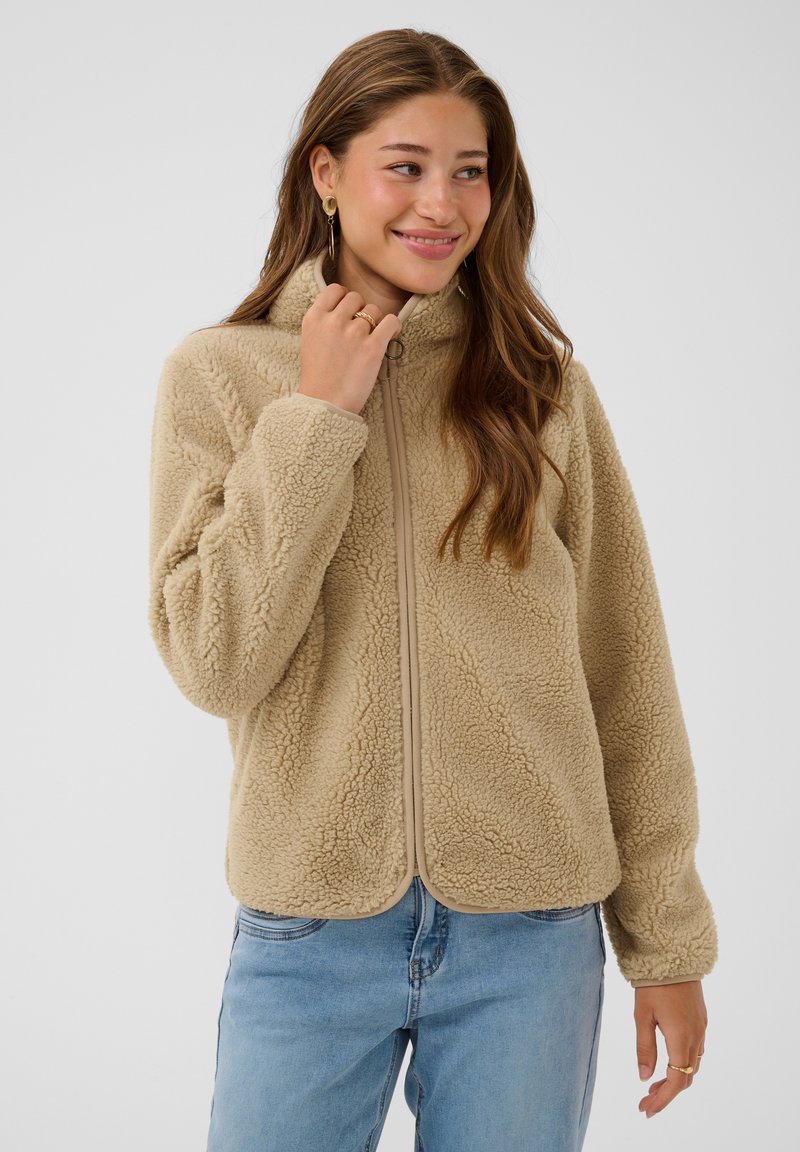 Young woman wearing beige fleece jacket and blue jeans, holding jacket collar and smiling, standing against plain background.