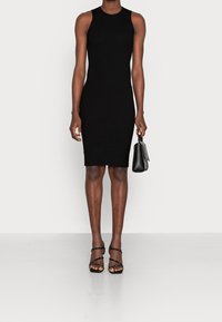 Robe noire en maille côtelée, sans manches, coupe près du corps et col rond. Accessoirisée d'un petit sac à main noir.