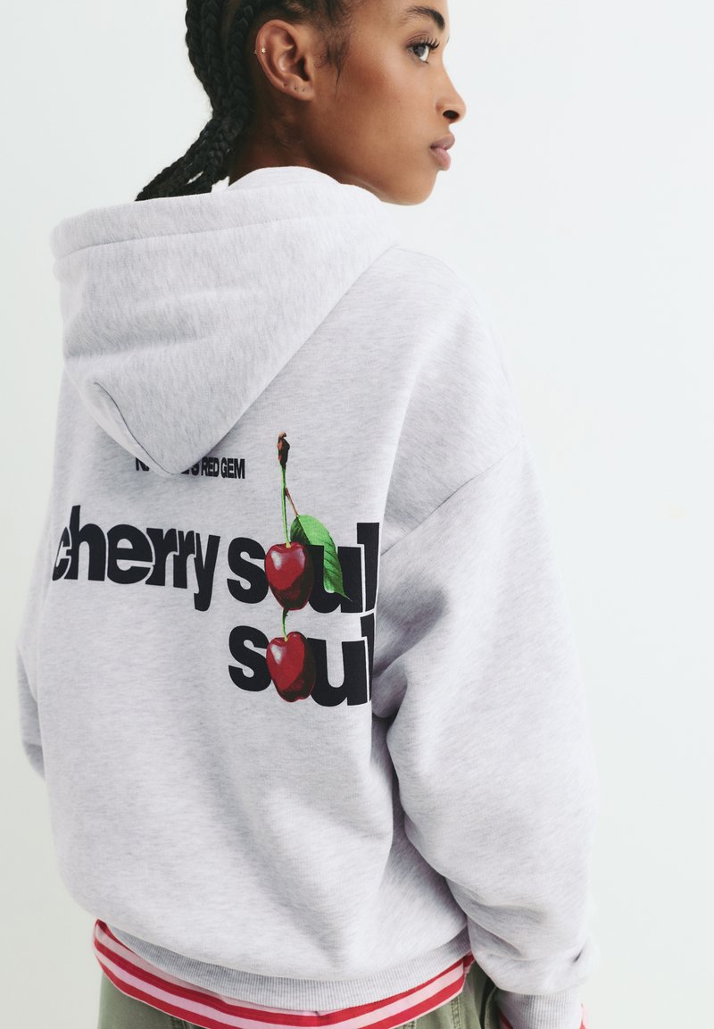 Felpa grigia con cappuccio e cordoncino, caratterizzata da testo nero e una grafica di due ciliegie sul retro. Il testo recita "cherry soul". Maniche lunghe.