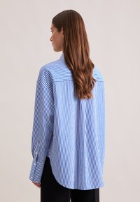 Camicia a righe blu e bianche con taglio dritto, polsini con bottoni e orlo curvo. Il materiale sembra leggero e ha una texture liscia.