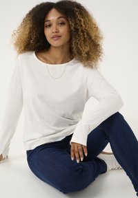Weißes Langarmoberteil aus einem weichen Stoff, kombiniert mit dunkelblauen Jeans. Auffällige goldene Halskette und hellfarbige Sneakers. Modell sitzt.